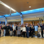 Eine große Gruppe von Menschen steht gemeinsam in einem großen Raum und lächelt in die Kamera.