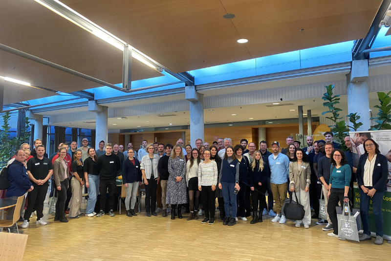 Eine große Gruppe von Menschen steht gemeinsam in einem großen Raum und lächelt in die Kamera.